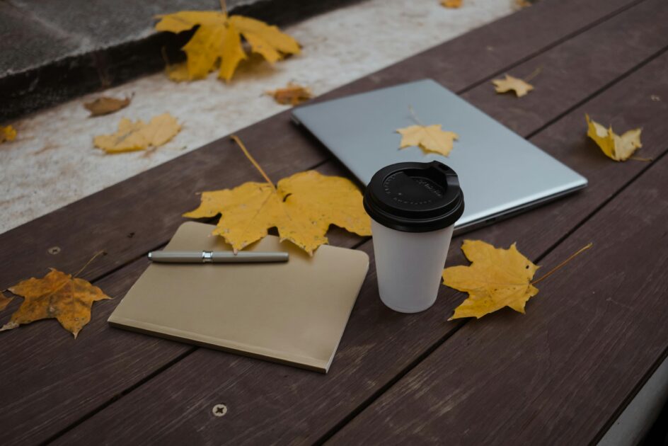 Autumn scene with a coffee cup, laptop, and notebook on a wooden bench surrounded by fall leaves.